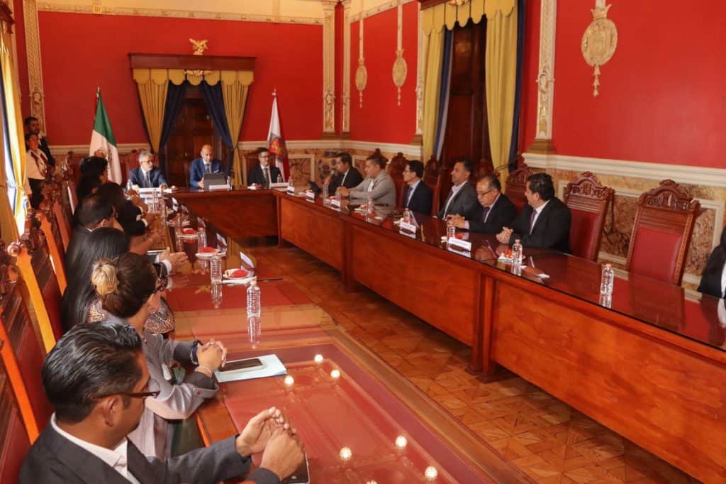 Acuerdo formal en una reunión gubernamental con funcionarios en una sala con paredes rojas y cortinas doradas.