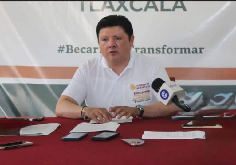 Joven hombre sentado en una mesa con micrófono, en un evento de Tlaxcala durante una conferencia de prensa o reunión oficial.