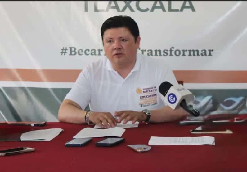 Joven hombre sentado en una mesa con micrófono, en un evento de Tlaxcala durante una conferencia de prensa o reunión oficial.