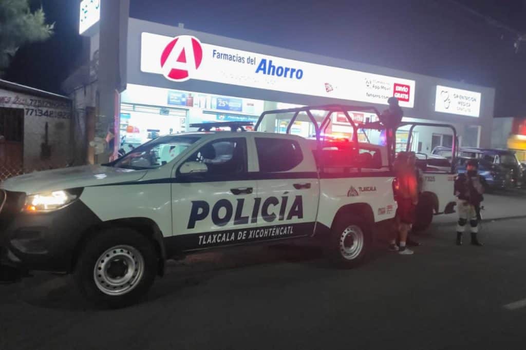 Policía de Tlaxcala en una patrulla en la calle frente a una farmacia del ahorro durante la noche, con luces rojas y personas cerca del vehículo.