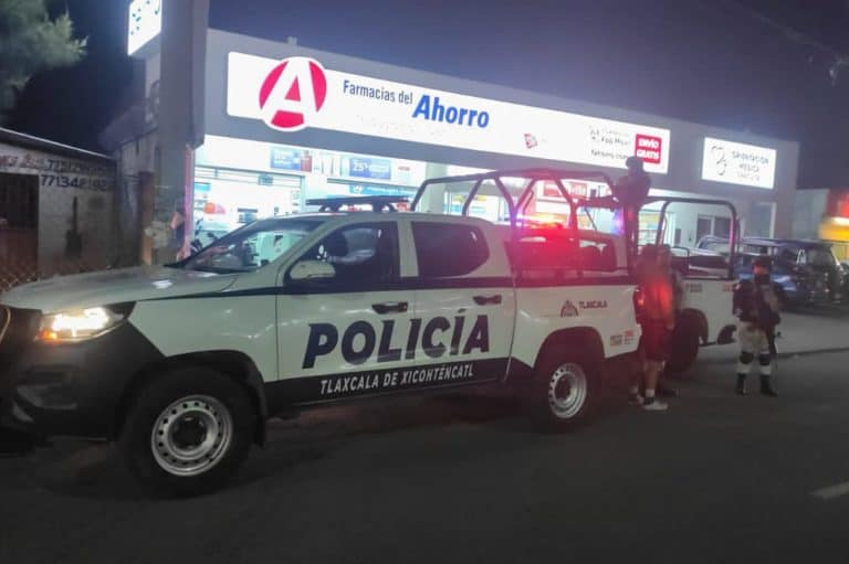 Policía de Tlaxcala en una patrulla en la calle frente a una farmacia del ahorro durante la noche, con luces rojas y personas cerca del vehículo.