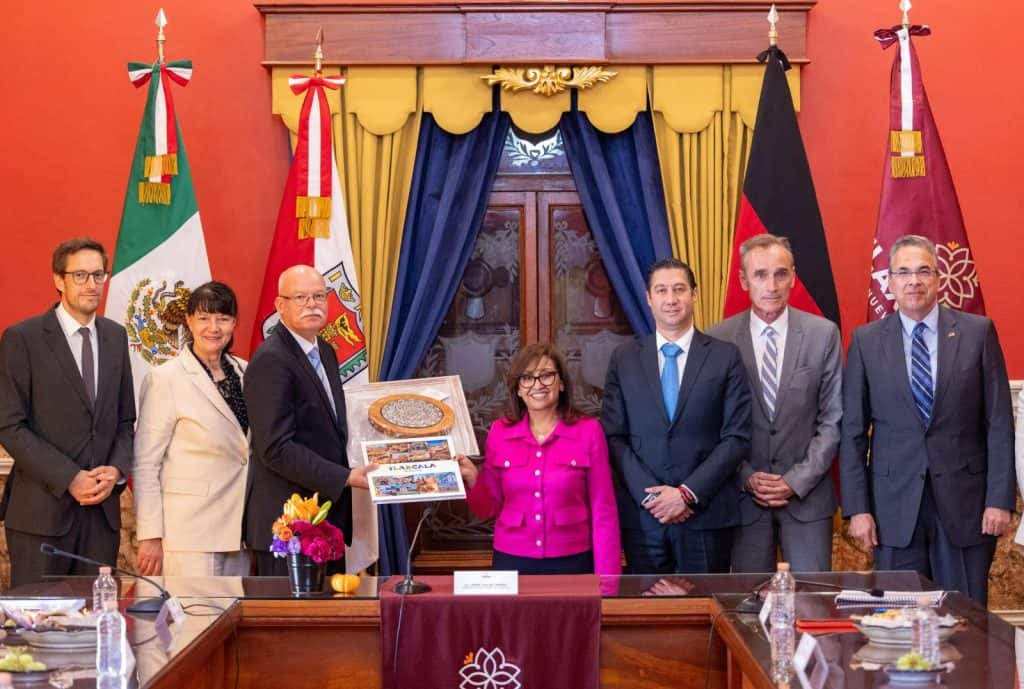 Funcionarios y líderes políticos en una reunión oficial sosteniendo un reconocimiento en un fondo con banderas de México, Tlaxcala, Alemania, y una sala decorada con cortinas azules y doradas.