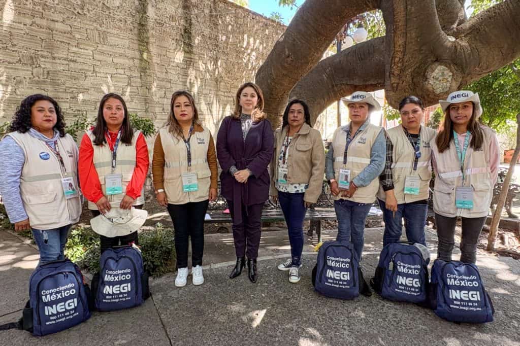 Personal de INEGI en una actividad al aire libre frente a un árbol grande, con bultos y gafetes de identificación, participando en una tarea relacionada con censos o encuestas.