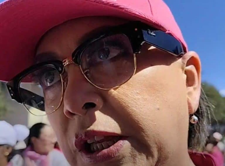 Mujer con gafas oscuras y sombrero rosa en un evento al aire libre, con expresión de preocupación o reflexión.