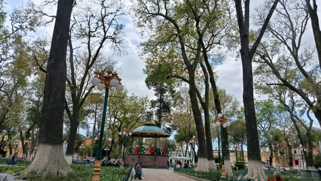 Árboles grandes en un parque con faroles antiguos y carrusel en el centro, bajo un cielo nublado, en Tlaxcala, México, creando un ambiente tranquilo y pintoresco.