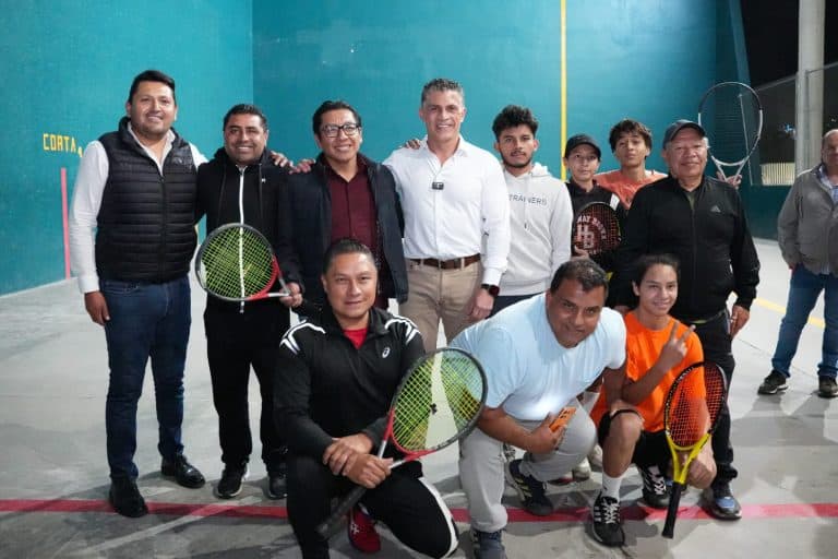 Jugadores y entrenadores de tenis de mesa en una cancha en interiores, algunos sosteniendo raquetas y haciendo el signo de paz, con rostros sonrientes, en medio de un evento deportivo en Tlaxcala, México.