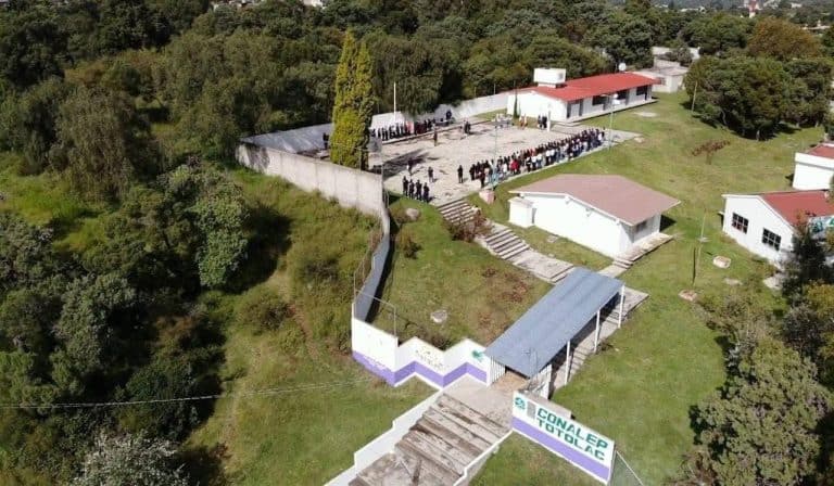 Estudiantes y docentes en una reunión al aire libre en un colegio de Tlaxcala, rodeado de áreas verdes y árboles, con varias edificaciones y una valla que lleva el nombre del plantel.