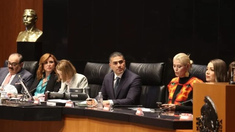 Busto de perfil de un hombre en mármol en la esquina izquierda de la imagen, fondo en tonos oscuros y mesa de reunión con varias personas sentadas con atención, algunas con documentos y teléfonos móviles, en un entorno formal y gubernamental.