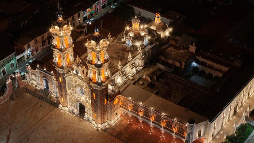 Imágen aérea de una iglesia iluminada por la noche en un entorno urbano en Tlaxcala, México. La iglesia presenta arquitectura colonial con torres y cúpulas decoradas con iluminación cálida y destacada, resaltando en el cielo nocturno.