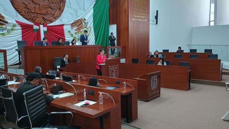 Juez en una sala de audiencias del Congreso de Tlaxcala durante una sesión de discusión y votación. El salón está decorado con la bandera y el escudo de Tlaxcala, con varias personas participando en el proceso legislativo.