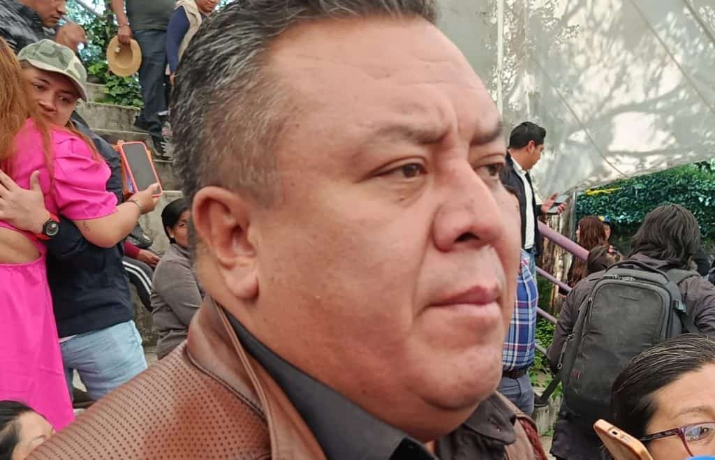 Hombre con expresión seria en medio de un evento con varias personas, algunos usando mochilas y otros tomando fotografías, en un entorno al aire libre con techos de plástico y vegetación.