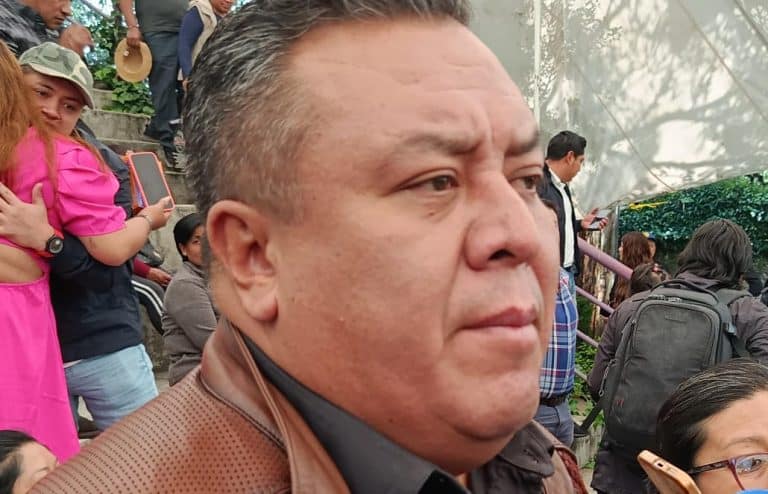 Hombre con expresión seria en medio de un evento con varias personas, algunos usando mochilas y otros tomando fotografías, en un entorno al aire libre con techos de plástico y vegetación.