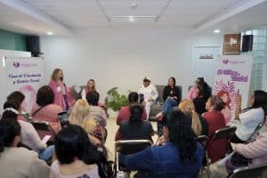 Mujer en vestido rosa hablando en panel de discusión con otras mujeres al fondo, en un evento de sensibilización sobre la violencia de género, en Tlaxcala, México.