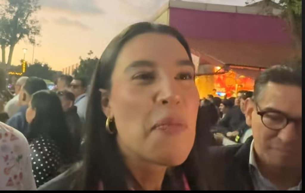 María Acosta en un evento público, rodeada de muchas personas, en un entorno al aire libre durante el atardecer, mostrando una expresión seria y segura, con fondo ilustrando actividad social.