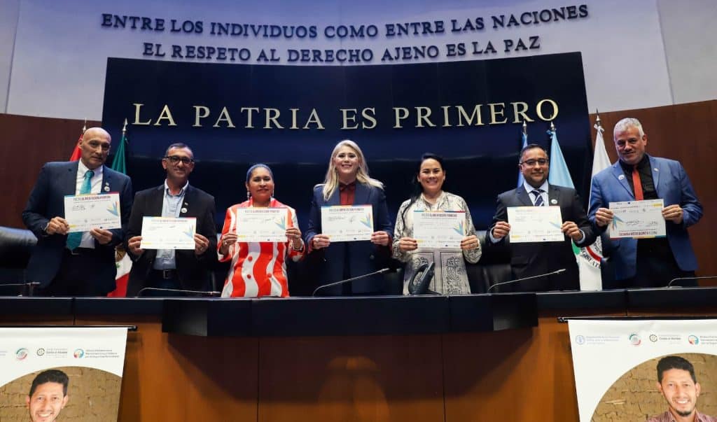 Diplomáticos de diferentes sectores sosteniendo certificados en un acto oficial en un auditorio.