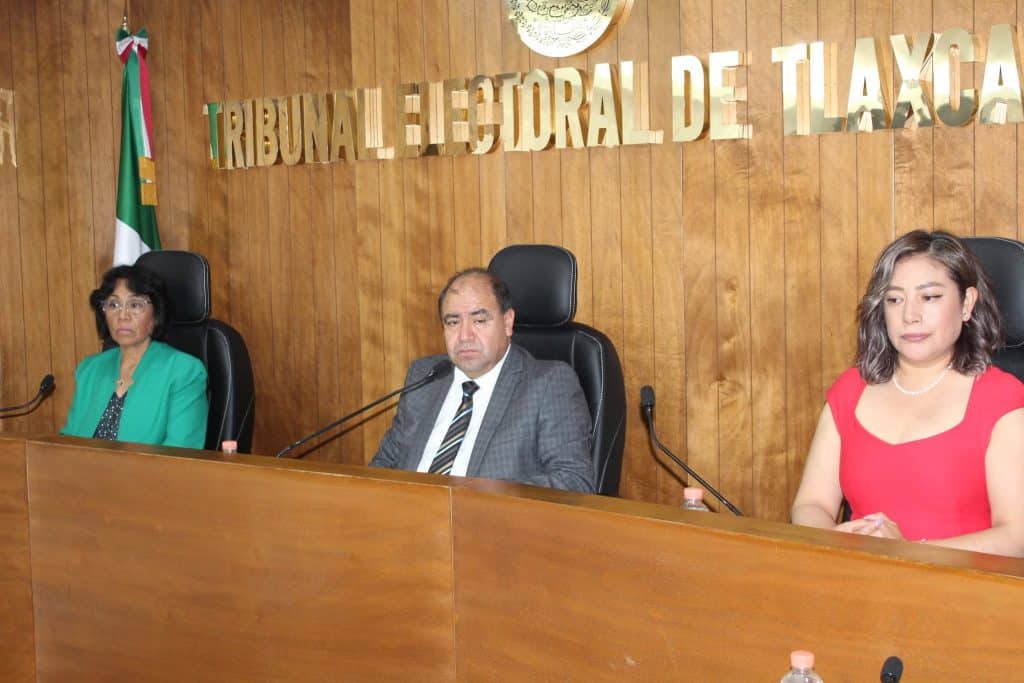 Jueces del Tribunal Electoral de Tlaxcala en una sesión formal, sentados en un panel con fondo de madera y la bandera de México a la izquierda.