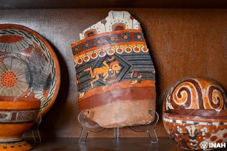 Pieza de cerámica prehispánica con decoraciones en colores naranja, negro y blanco, que representa un animal en un entorno con motivos geométricos, exhibida en un soporte de exhibición.