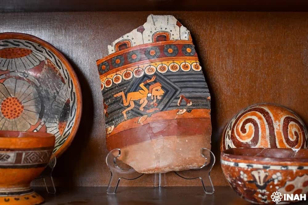 Pieza de cerámica prehispánica con decoraciones en colores naranja, negro y blanco, que representa un animal en un entorno con motivos geométricos, exhibida en un soporte de exhibición.