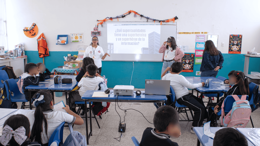 Niñas y niños en un aula escolar en clase, con dos maestras frente a la pizarra, en una imagen que muestra un ambiente educativo y de aprendizaje.