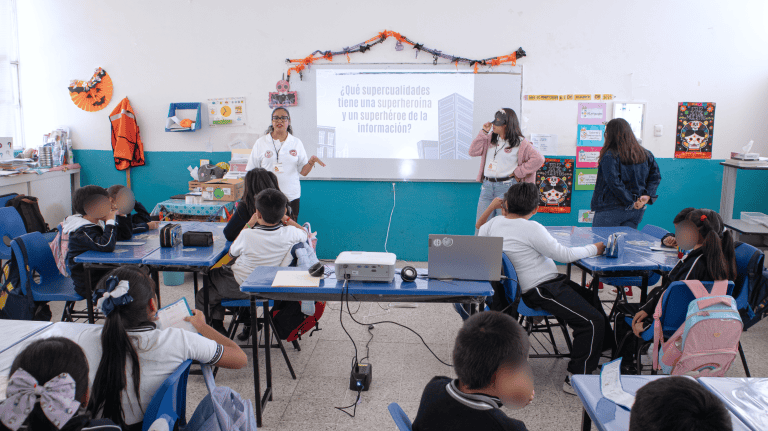 Niñas y niños en un aula escolar en clase, con dos maestras frente a la pizarra, en una imagen que muestra un ambiente educativo y de aprendizaje.
