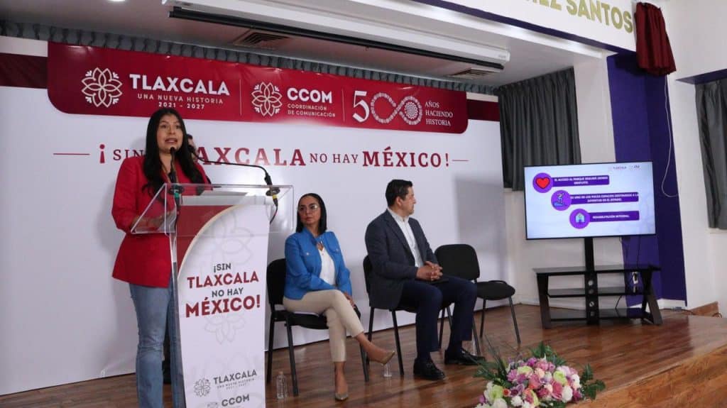 Alto en la imagen, una mujer con blazer rojo habla en un micrófono en una conferencia o evento oficial en Tlaxcala, con un cartel y un monitor a su lado.