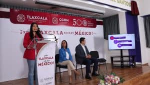Alto en la imagen, una mujer con blazer rojo habla en un micrófono en una conferencia o evento oficial en Tlaxcala, con un cartel y un monitor a su lado.