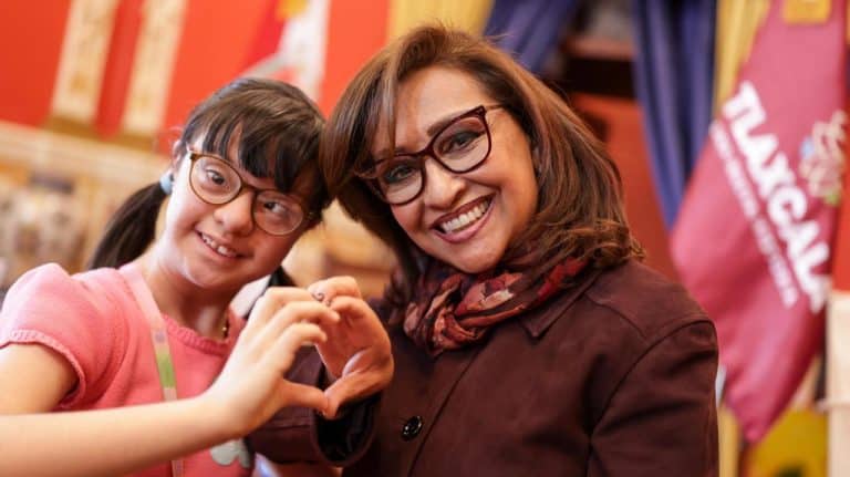Sonrientes mujer y niña haciendo un corazón con sus manos en un evento, representando alegría y unión familiar en un entorno cultural y educativo en Tlaxcala, México.