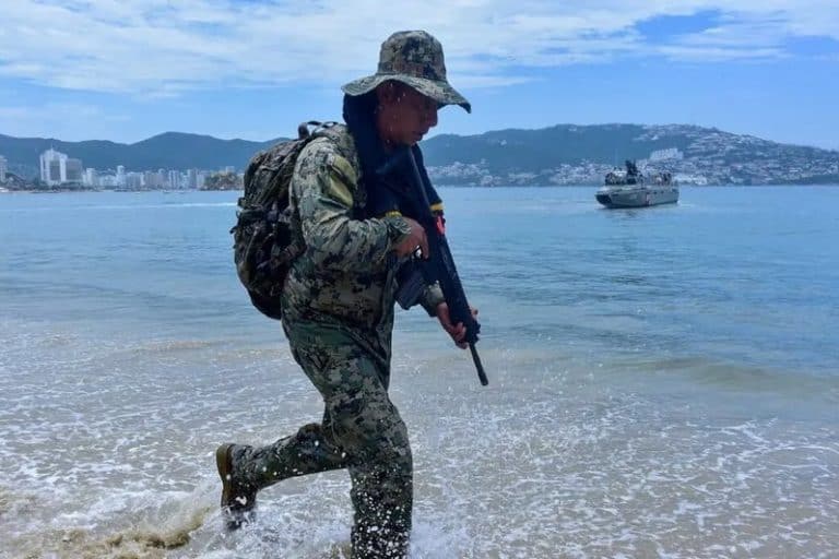 Soldado en la playa con uniforme de camuflaje, portando un fusil, caminando hacia el agua en una zona costera con ciudad y barcos de fondo.