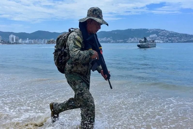 Soldado en la playa con uniforme de camuflaje, portando un fusil, caminando hacia el agua en una zona costera con ciudad y barcos de fondo.