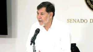 Diputado en conferencia en el Senado de México, vestido de blanco y con barba gris, durante una reunión oficial, frente a un fondo con logotipo y la palabra "SENADO".