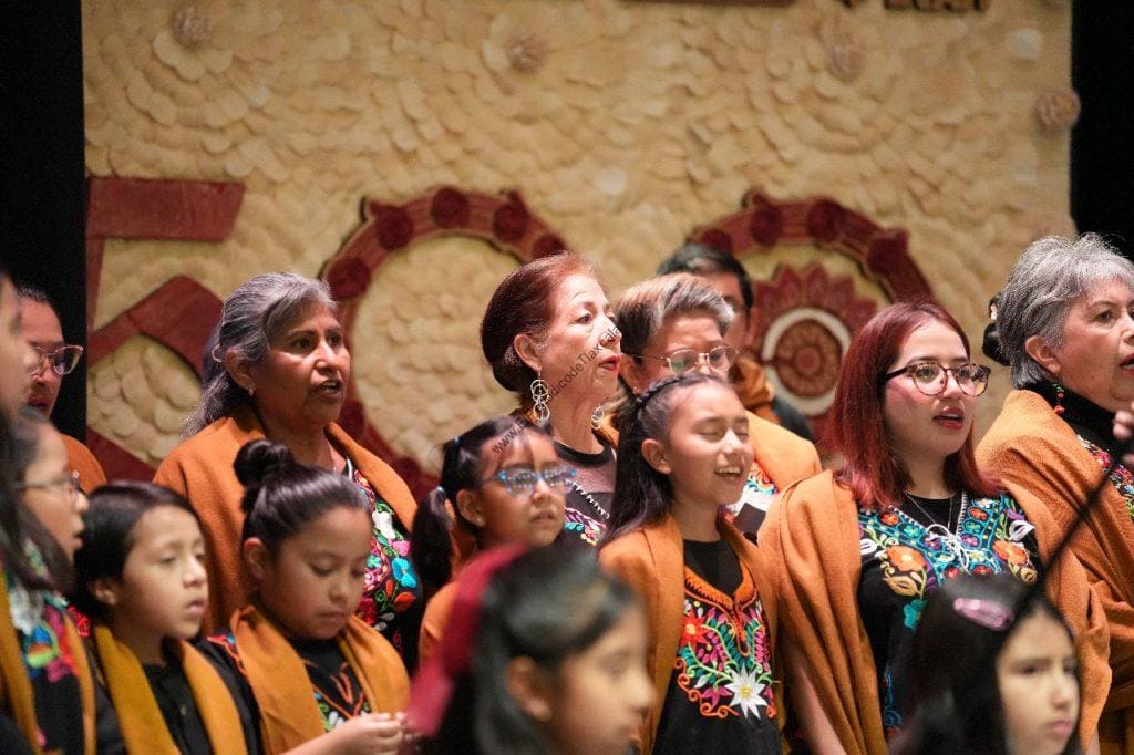 Vestido tradicional con bordados coloridos en un grupo diverso de mujeres y niñas mexicanas, participando en un evento cultural o celebración en un entorno con decoración artesanal de fondo.