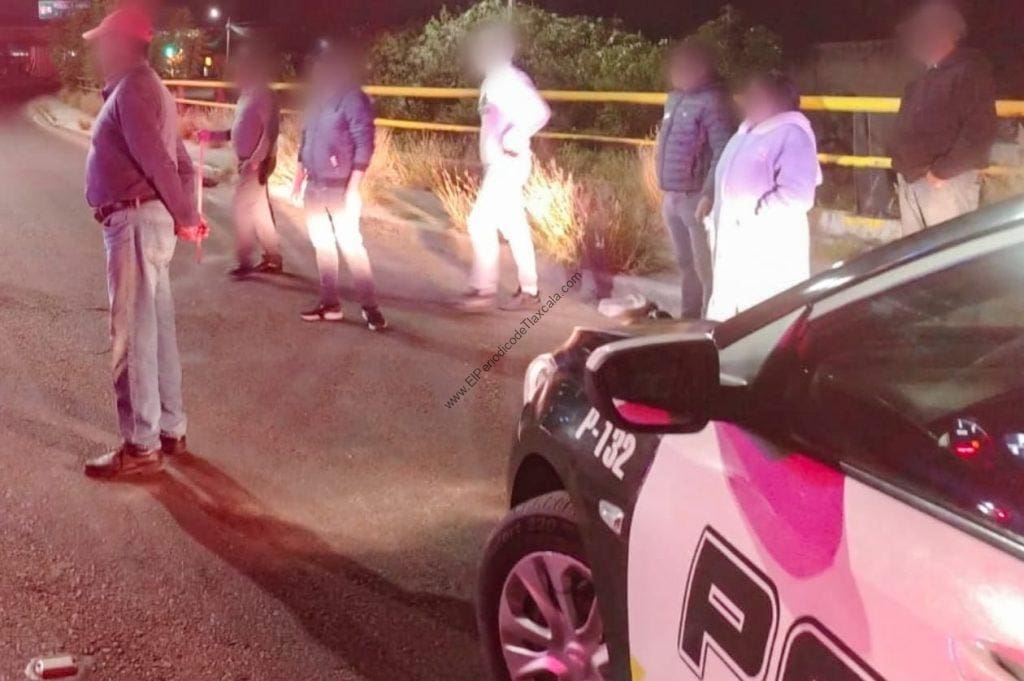 Difuntos en la escena con oficiales y personas observando en la noche, junto a un vehículo de policía en Tlaxcala, México. La imagen muestra una situación de emergencia o investigación en un lugar oscuro, en la calle.