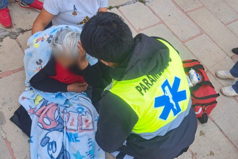 Persona de la tercera edad recibiendo atención médica de un paramédico en la calle. La mujer está acostada sobre una cobija con estampados infantiles, mientras el paramédico la atiende con cuidado.