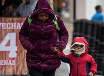 Niña y mujer con ropa de abrigo en la calle, ambas usando mascarillas y abrigos rojos y morados, sosteniéndose de la mano, en un día frío y nublado en Tlaxcala, México.