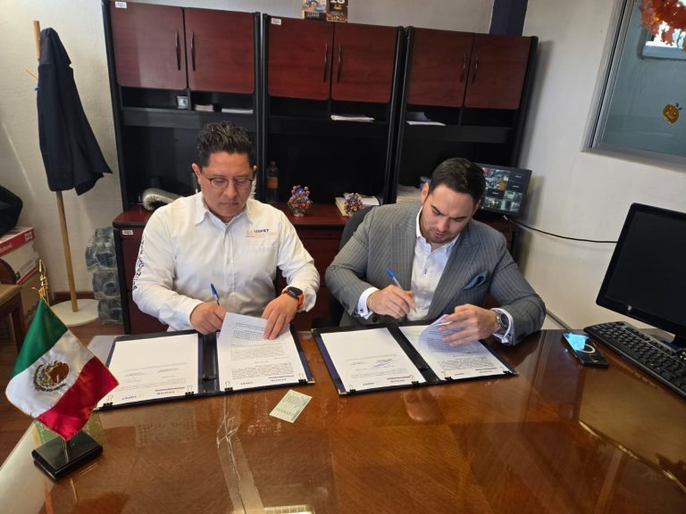 Firma de dos hombres en un escritorio, firmando documentos oficiales, con bandera de México en primer plano y fondo de estanterías y equipo de oficina.