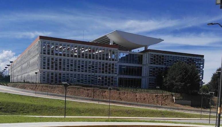 Edificio moderno con diseño arquitectónico innovador y amplias ventanas, ubicado en un entorno urbano con césped y caminos peatonales en primer plano, bajo un cielo parcialmente nublado.