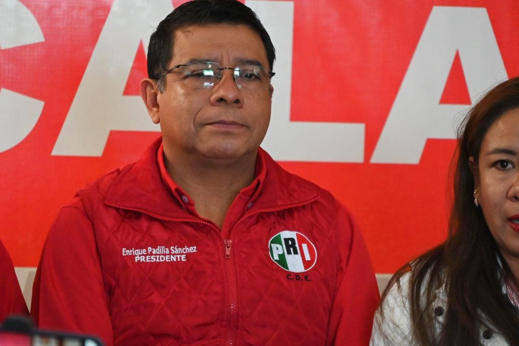 Camisa roja con logotipo del PRI y texto que indica "Enrique Padilla Sánchez, PRESIDENTE". Hombre con gafas en una conferencia de prensa, con fondo rojo y letras blancas del partido PRI.