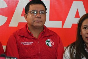 Camisa roja con logotipo del PRI y texto que indica "Enrique Padilla Sánchez, PRESIDENTE". Hombre con gafas en una conferencia de prensa, con fondo rojo y letras blancas del partido PRI.