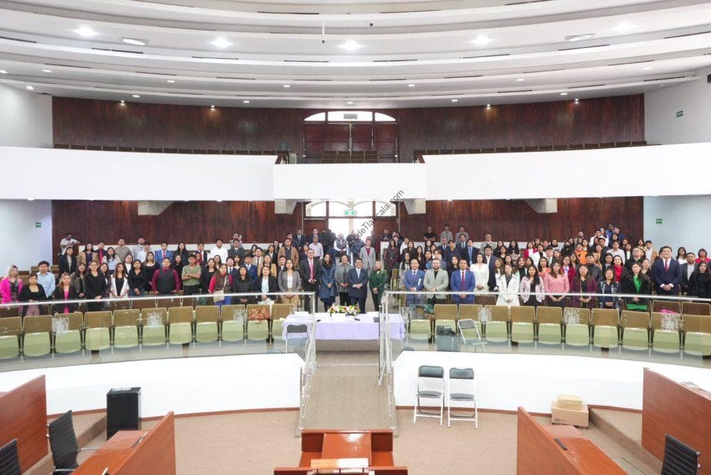 Amanecer en el acto solemne con un grupo diverso de personas, vestidas formalmente, en un auditorio de arquitectura moderna con paredes de madera y amplio espacio, en Tlaxcala, México.