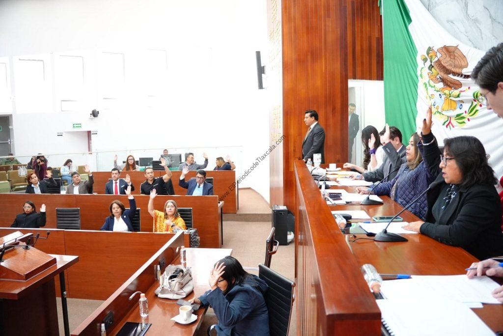 Jurado en sesión en un parlamento, con varias personas levantando la mano durante una votación, en un entorno formal y con bandera de México en el fondo.
