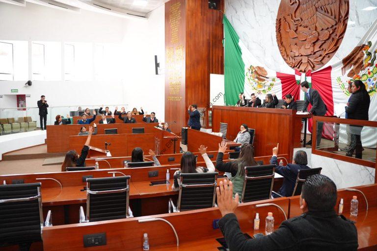 Diputados en sesión en el Congreso de Tlaxcala, algunos levantan la mano, otros están votando, con banderas de México y el escudo nacional en el fondo.