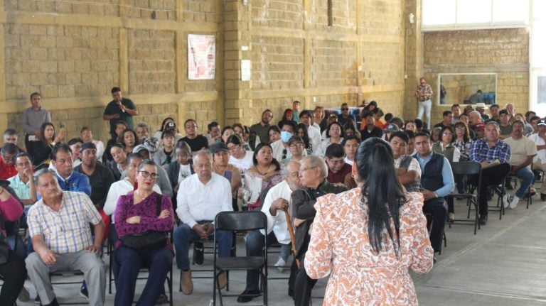 Asiste un gran número de personas a una conferencia o evento en un espacio cerrado con paredes de ladrillo y ventanas grandes, sentado en sillas, en un ambiente formal y de interés comunitario.