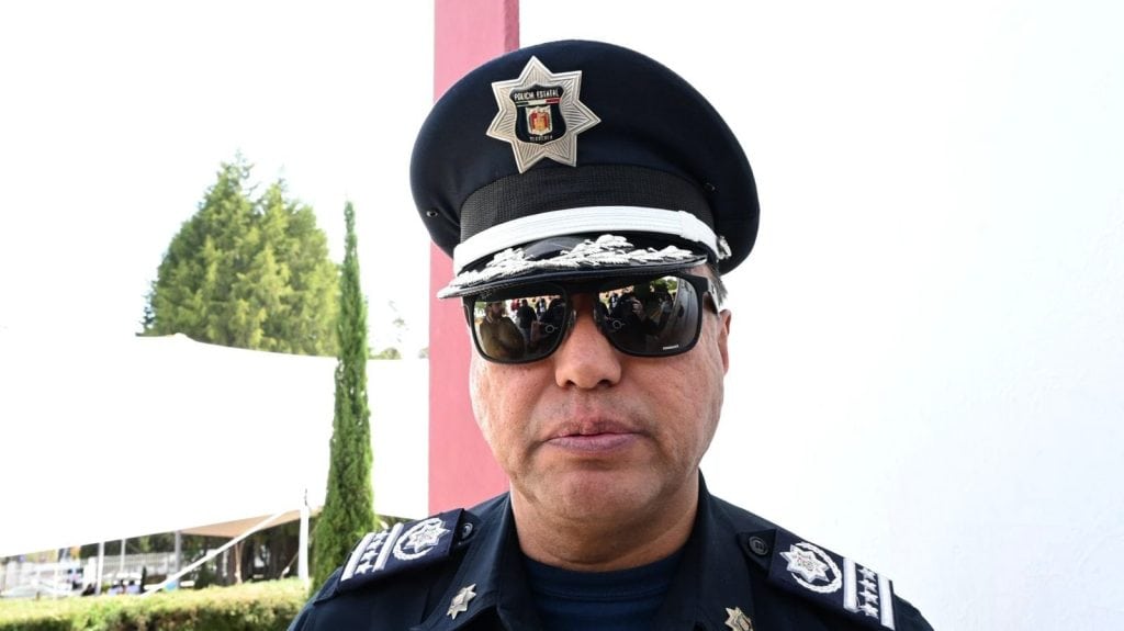 Policía estatal de Tlaxcala con uniforme y gorra, usando gafas oscuras, en un día soleado, en un entorno con árboles y una estructura en el fondo.