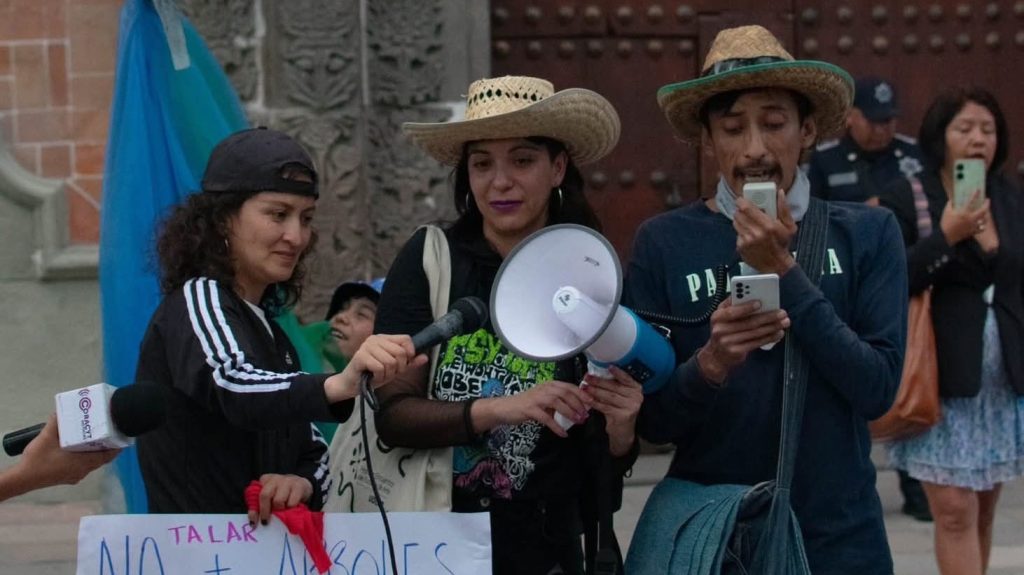 Abogada con sombrero y megáfono participando en una protesta en Tlaxcala, México, junto a otras personas que también usan sombreros y sostienen teléfonos móviles en una manifestación pública.