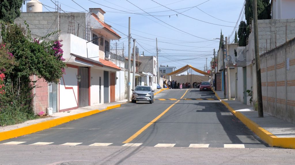 Calle en Tlaxcala con casas y redes eléctricas, un coche estacionado y una carpa en la distancia, ambiente urbano tranquilo y residencial, día soleado con cielo despejado.