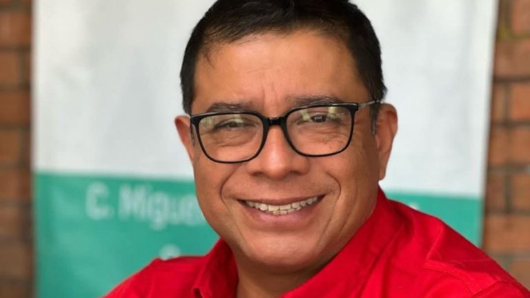 Alté text: Fotografía de un hombre con gafas y camisa roja sonriendo, en un fondo difuso con paredes de ladrillo y cartel.