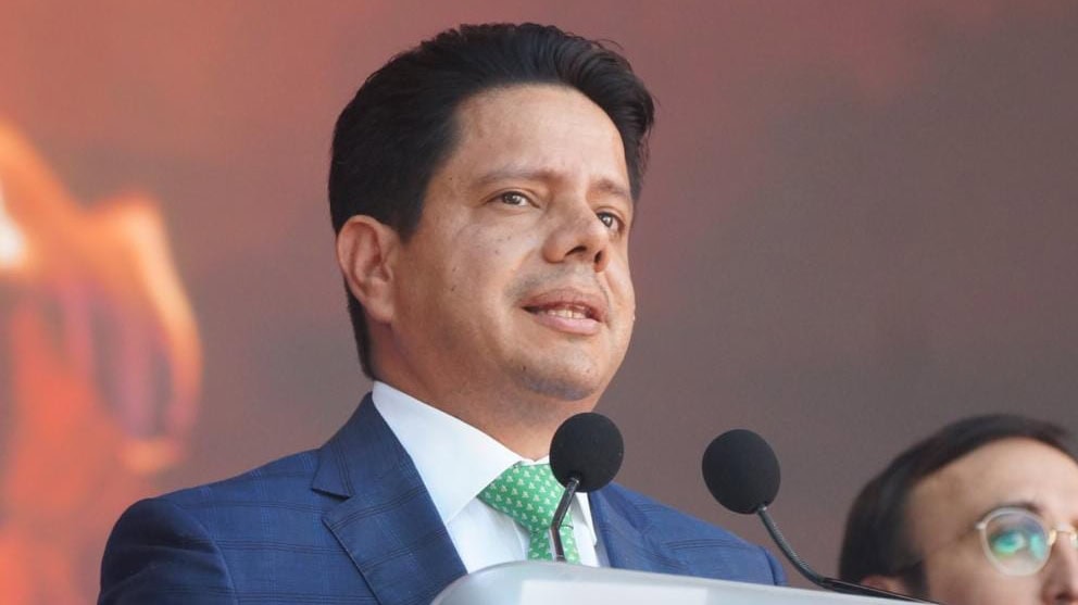 Conferencia de prensa de un político en un evento formal, vestido con traje azul y corbata verde, hablando frente a micrófonos en un escenario con fondo difuso y cálido, en un contexto de noticias.