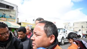 Político en entrevista en calle de Tlaxcala, rodeado de varias personas y coches, en un día nublado.