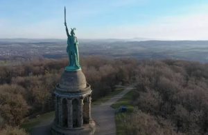 Gran estatua de la libertad en un parque rodeado de árboles y caminos, con un cielo despejado en un día soleado.