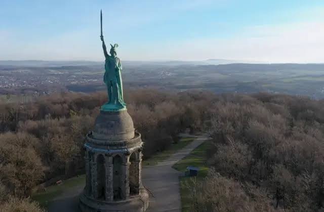 Gran estatua de la libertad en un parque rodeado de árboles y caminos, con un cielo despejado en un día soleado.